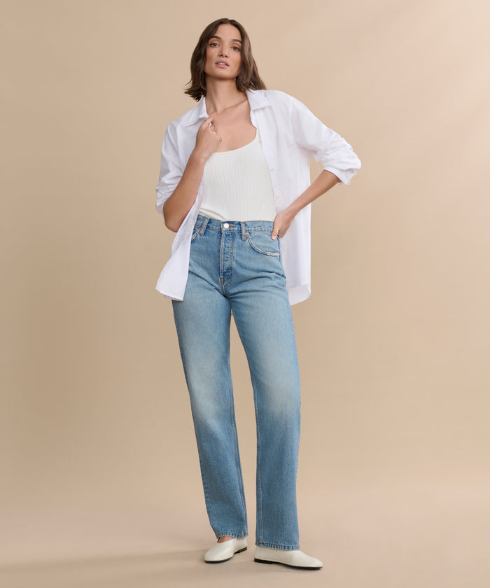 A woman poses against a beige background wearing a white open button-up shirt over a tank top, paired with the 90s Jean in light blue and white shoes. One hand is in her pocket while the other touches her collar.