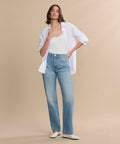 A woman poses against a beige background wearing a white open button-up shirt over a tank top, paired with the 90s Jean in light blue and white shoes. One hand is in her pocket while the other touches her collar.