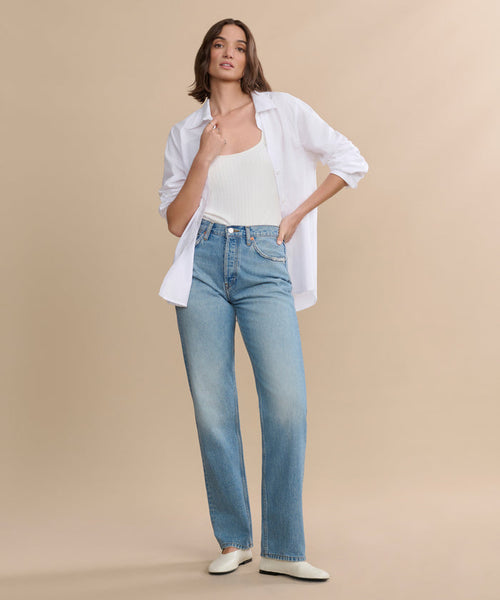 A woman poses against a beige background wearing a white open button-up shirt over a tank top, paired with the 90s Jean in light blue and white shoes. One hand is in her pocket while the other touches her collar.