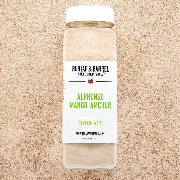 A jar of Alphonso Mango Amchur spice on a background of loose amchur with the label 'BURLAP & BARREL' and 'Alphonso Mango Amchur