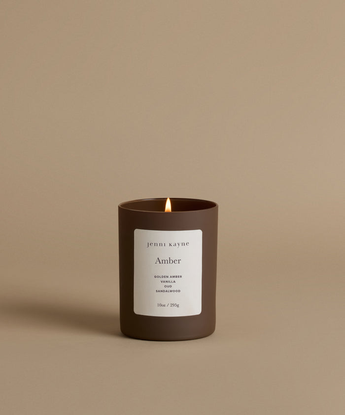 A lit candle in a matte dark brown glass jar with a white label reading 'jenni kayne' and 'Amber', set against a solid warm beige background.