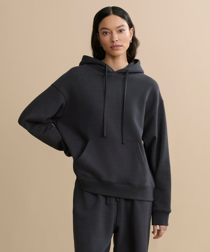 A person with long dark hair stands against a beige background, wearing the cozy washed black Bay Hoodie with a front pocket and matching black pants. One hand rests on their hip while the other hangs by their side.