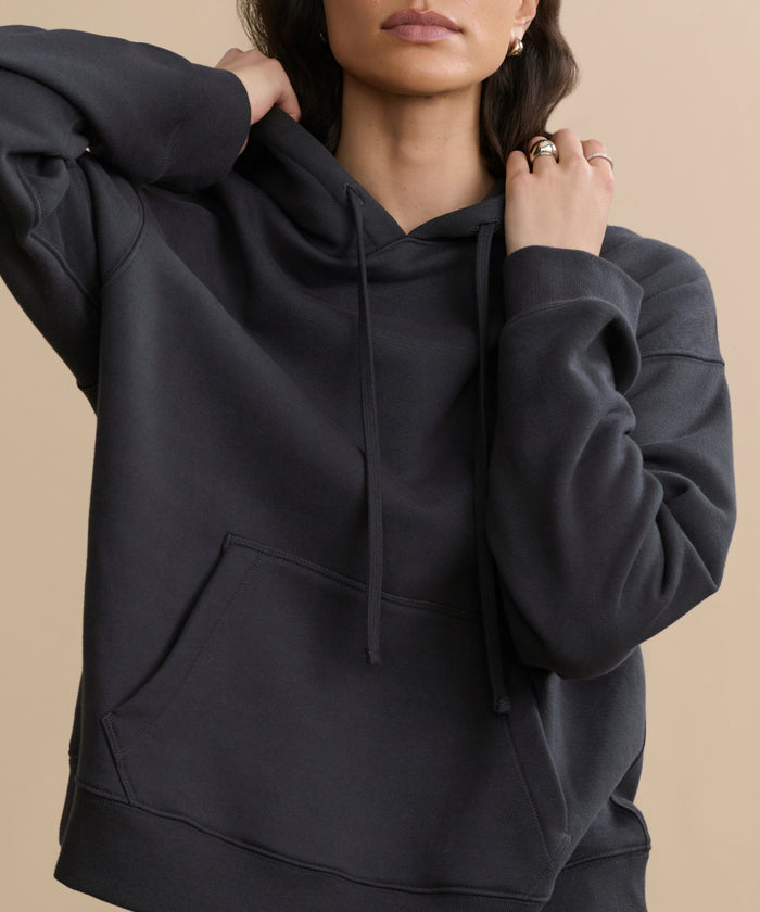 A woman adjusts the hood of the Bay Hoodie—a loose-fitting, washed black pullover with a front pocket and drawstrings—standing against a beige background. Her relaxed pose highlights the cozy and casual style of the hoodie.