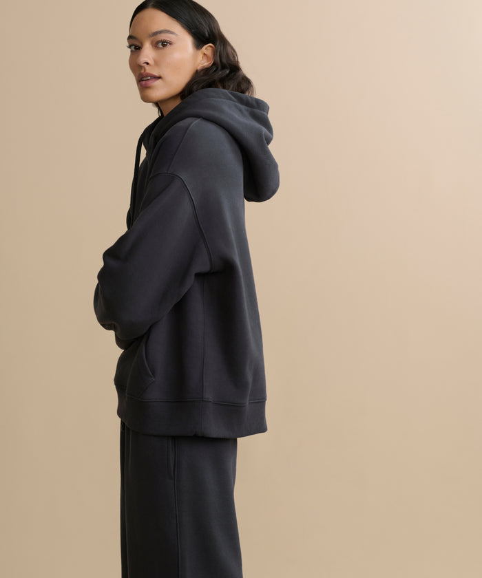 A person with long dark hair stands sideways, arms crossed, wearing the loose-fitting Bay Hoodie in washed black and matching pants against a plain beige background.