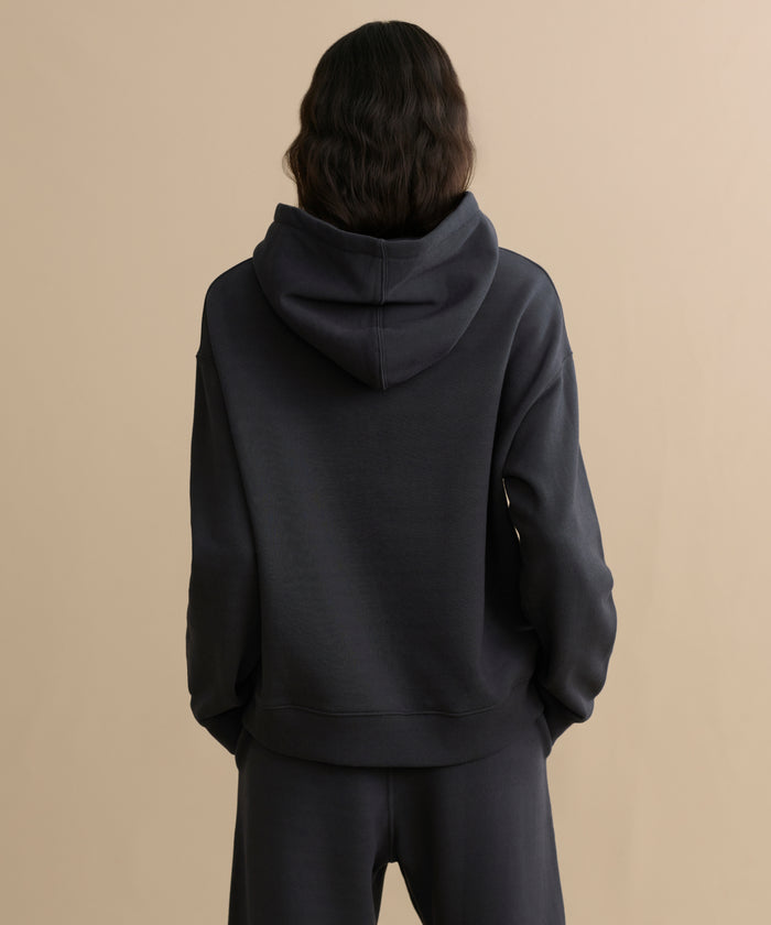 A person with long, wavy dark hair stands with their back to the camera, wearing the cozy Bay Hoodie in washed black and matching pants, set against a beige background.