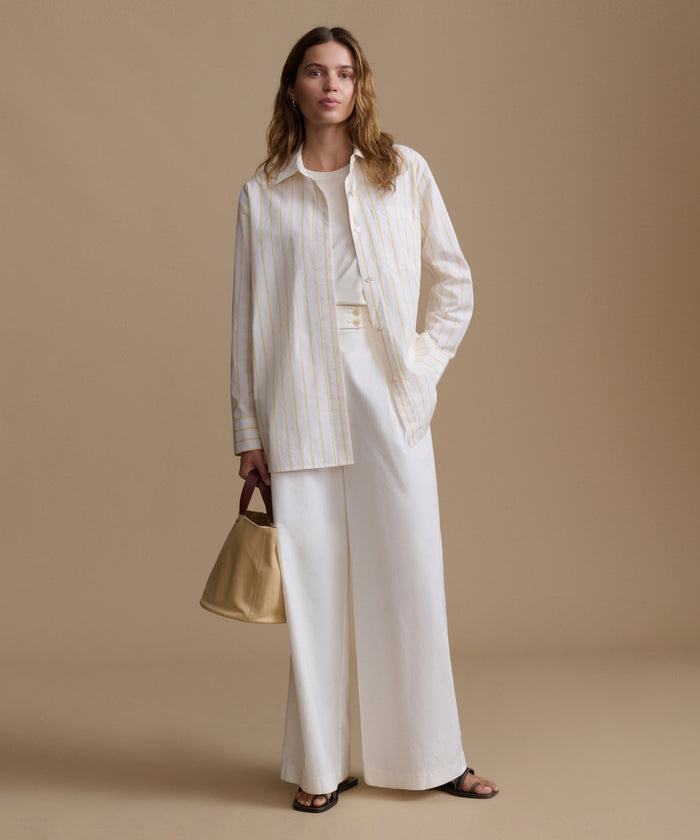Woman wearing a stripe button up shirt with white pants against a beige background.