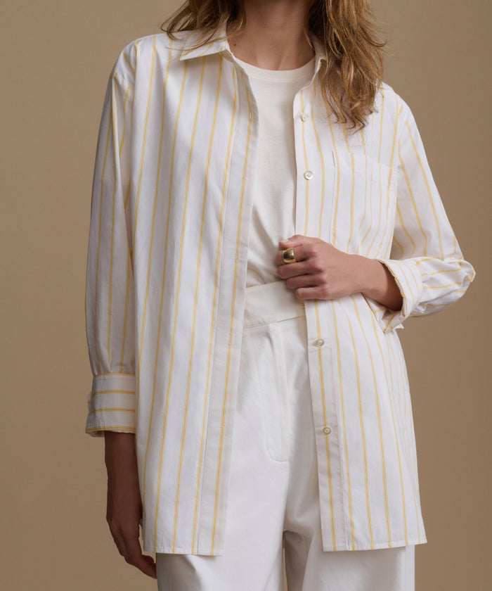 Woman wearing a stripe button up shirt with white pants against a beige background.