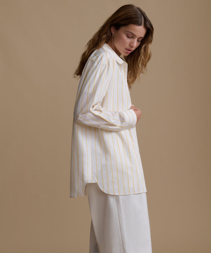 Woman wearing a stripe button up shirt with white pants against a beige background.