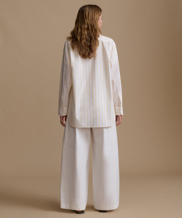 Woman wearing a stripe button up shirt with white pants against a beige background.