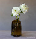 transparent brown hand-blown glass flower bud vase with white flowers on wood table and gray background