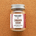 A glass jar of Cinnamon Verum spice with a label that reads 'Cinnamon Verum' and 'Zanzibar Archipelago, Tanzania'. The jar is placed against a textured background.