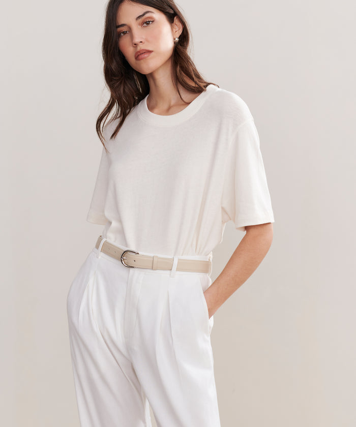 A woman with long brown hair stands against a plain light background wearing a loose white t-shirt tucked into high-waisted white pleated pants, styled with the Classic Belt made in Italy.