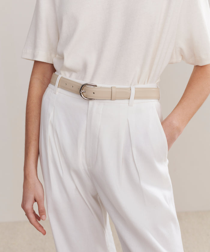 Person wearing a loose white t-shirt tucked into high-waisted white pleated trousers, accessorized with the Classic Belt. The minimal, monochrome outfit features their hand resting by their side.