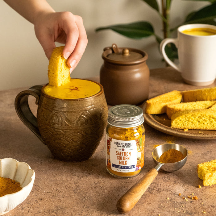 Person dipping a piece of biscotti into a mug with a latte made with Burlap & Barrel's Saffron Golden Milk  and biscotti pieces on a table.