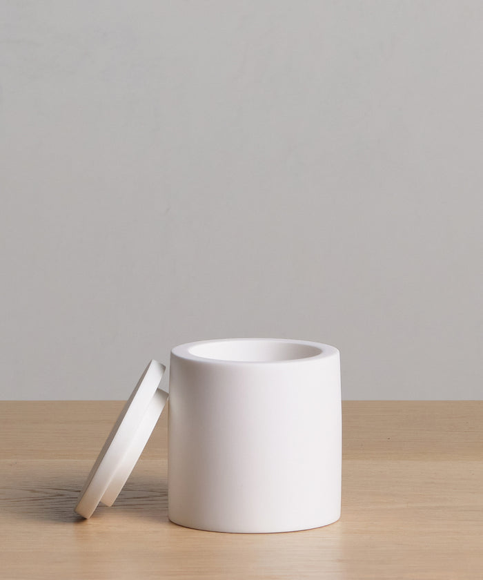 The Coast Canister is a minimalist, white cylindrical container with a matching lid, crafted from handcasted resin and displayed on a light wood surface against a plain light gray background.