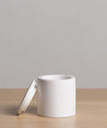 The Coast Canister is a minimalist, white cylindrical container with a matching lid, crafted from handcasted resin and displayed on a light wood surface against a plain light gray background.