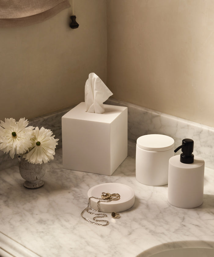 A marble countertop with white home accessories, featuring the Coast Canister, a matching tissue box, pump dispenser, jewelry dish, and a small vase with white daisies.