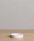 The Coast Soap Dish is a small, round, white resin dish, handcasted in Mexico. It sits on a light wooden surface with a plain, light gray background.