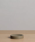 The Coast Soap Dish, a simple round and flat piece made from taupe handcast resin, rests on light wood with a plain light gray backdrop.