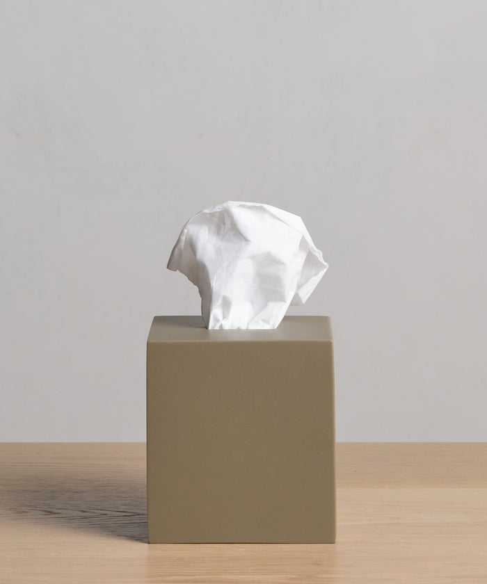 A beige Coast Tissue Cover, crafted from handcasted resin, rests on a light wooden surface with a crumpled white tissue partially pulled from the top against a plain light gray background.