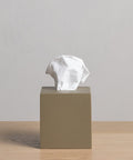 A beige Coast Tissue Cover, crafted from handcasted resin, rests on a light wooden surface with a crumpled white tissue partially pulled from the top against a plain light gray background.