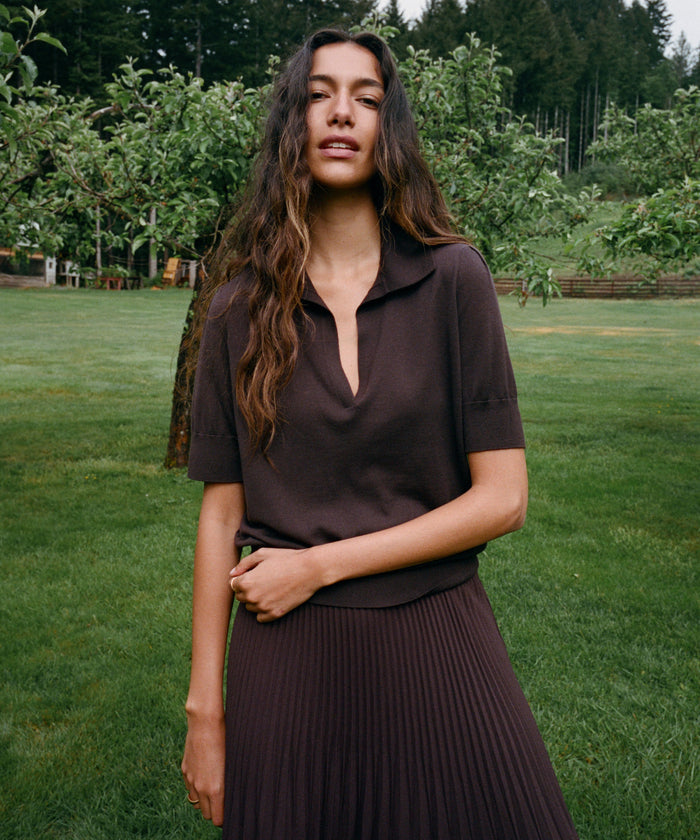 A woman standing outdoors on a grassy lawn wearing a dark brown short-sleeve collared top and matching pleated skirt, with trees in the background.