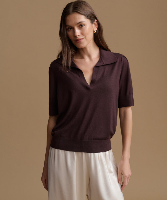A woman modeling a short-sleeve knit polo top in deep plum purple, paired with cream-colored pants, against a solid warm beige studio background.