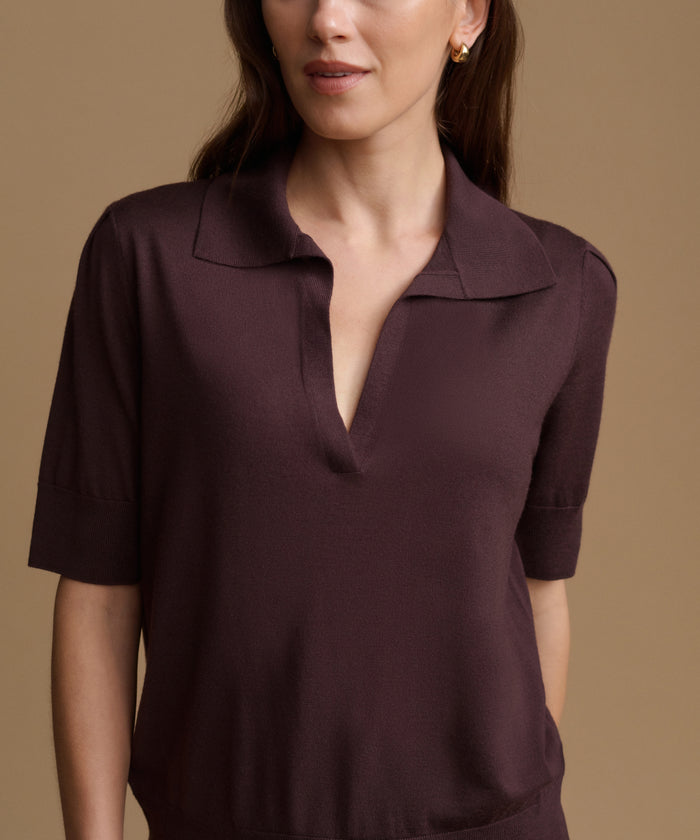 A close-up of a woman wearing a short-sleeve polo-style top in a deep plum or eggplant purple color, set against a solid warm beige background.