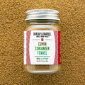 A glass jar of Burlap & Barrel Cumin Coriander Fennel spice mix, featuring a white label with red patterns and a silver metal lid, set against a textured background of loose ground coriander seeds.