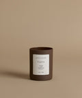A glass candle in th scent Cypress in a brown vessel against a beige background.