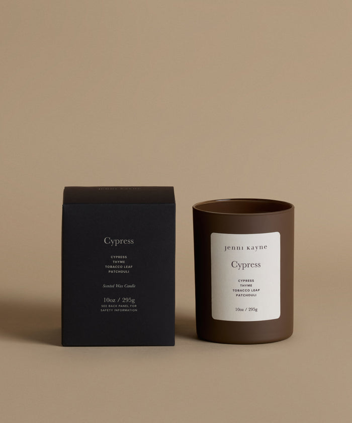 A candle in a dark brown glass jar with a white label next to its black packaging box, both featuring the brand name 'Jenii Kayne' and the scent 'Cypress', set against a solid beige background.