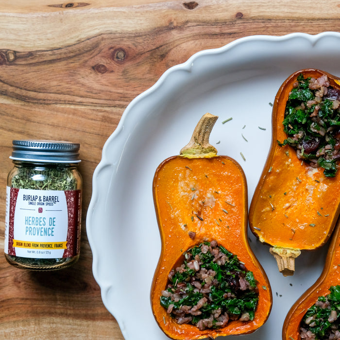 Butternut squash halves with stuffing on a white plate next to a jar of herbs de Provence on a wooden surface.