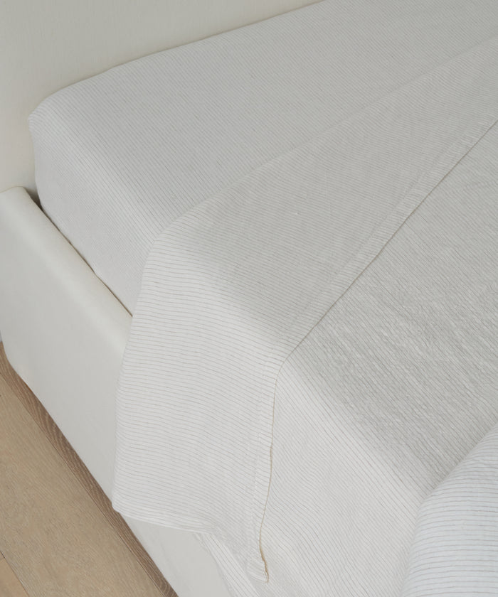 A neatly made bed features the Dusk Linen Flat Sheet, boasting a subtle flax stripe texture, paired with a white fitted sheet on a light-colored frame beside a wooden floor.
