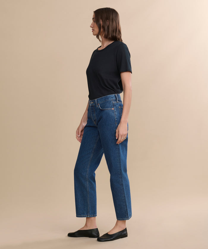 A woman in profile stands against a plain beige background, wearing a black short-sleeve shirt, blue Easy Straight Crop jeans with a relaxed leg, and black flats.