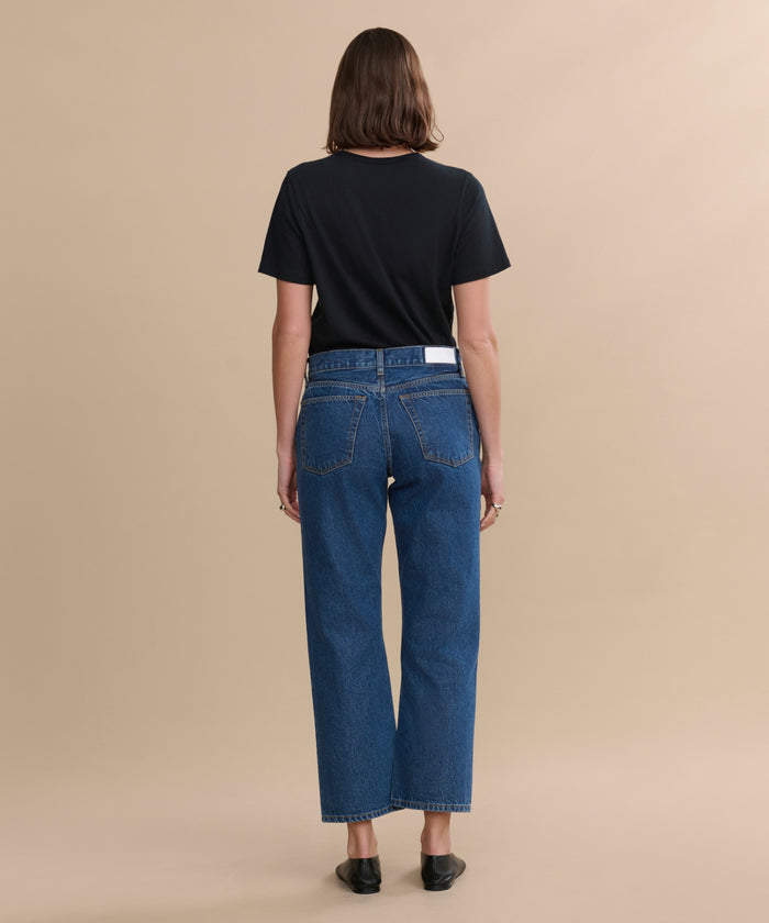 A person with straight brown hair faces away, wearing a black t-shirt, black shoes, and blue RE/DONE Easy Straight Crop jeans with a relaxed leg, against a plain beige background.
