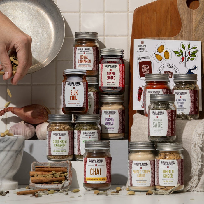 Jars of Burlap & Barrel spices and seasonings on a kitchen counter with a person preparing food.