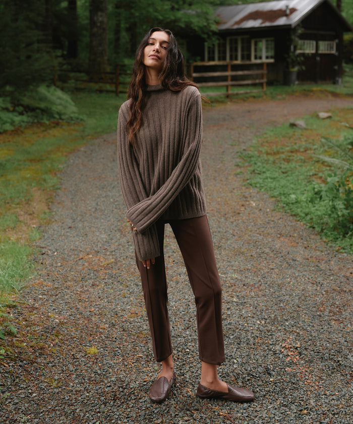 A woman stands on a gravel path in a wooded outdoor setting, wearing a ribbed brown mock-neck sweater, brown pants, and brown loafers.