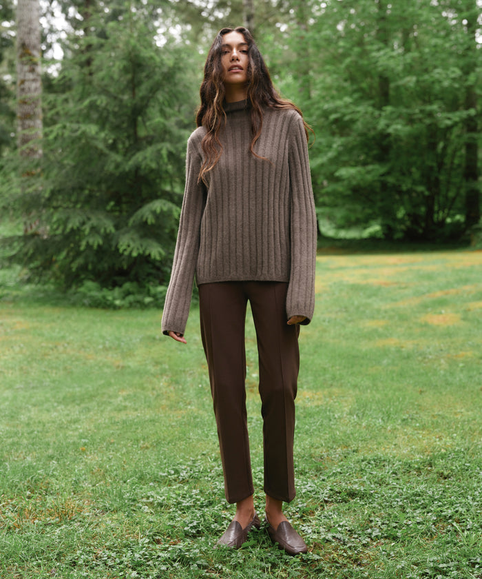 A woman stands outdoors in a grassy park area with trees in the background, wearing a ribbed brown turtleneck sweater, dark brown pants, and brown loafers.