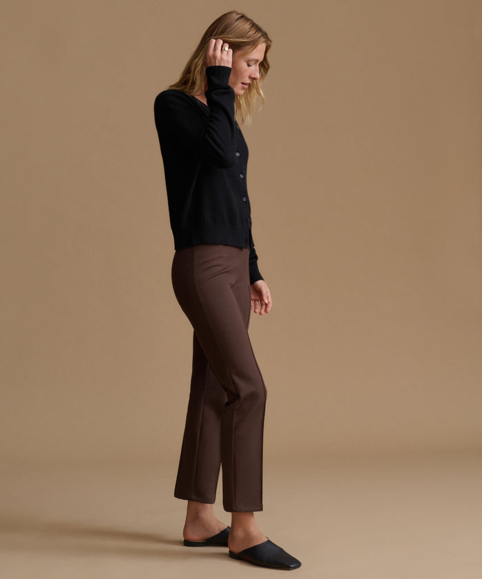 A woman stands in profile wearing a black long-sleeve button-front top, brown cropped pants, and black slip-on shoes against a plain warm beige studio background.