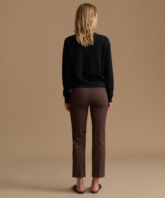 A woman viewed from behind wearing a black long-sleeve top and brown straight-leg trousers, standing against a solid beige studio background.