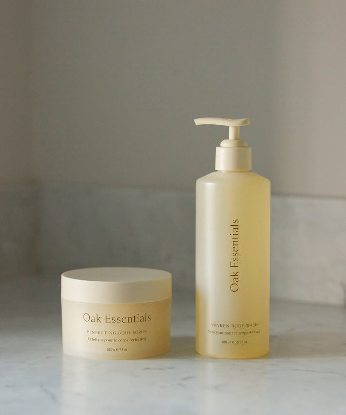 The Fresh Start Set, featuring a round jar of Perfecting Body Scrub and a tall pump bottle of Awaken Body Wash in minimalist beige packaging, sits on a marble surface—ideal for exfoliating and cleansing.