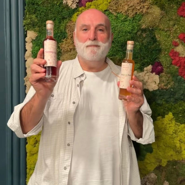 A man with a white beard and bald head holds up two bottles of syrup, one in each hand, in front of a green moss wall. He is wearing a white button-down shirt over a white t-shirt and black pants.