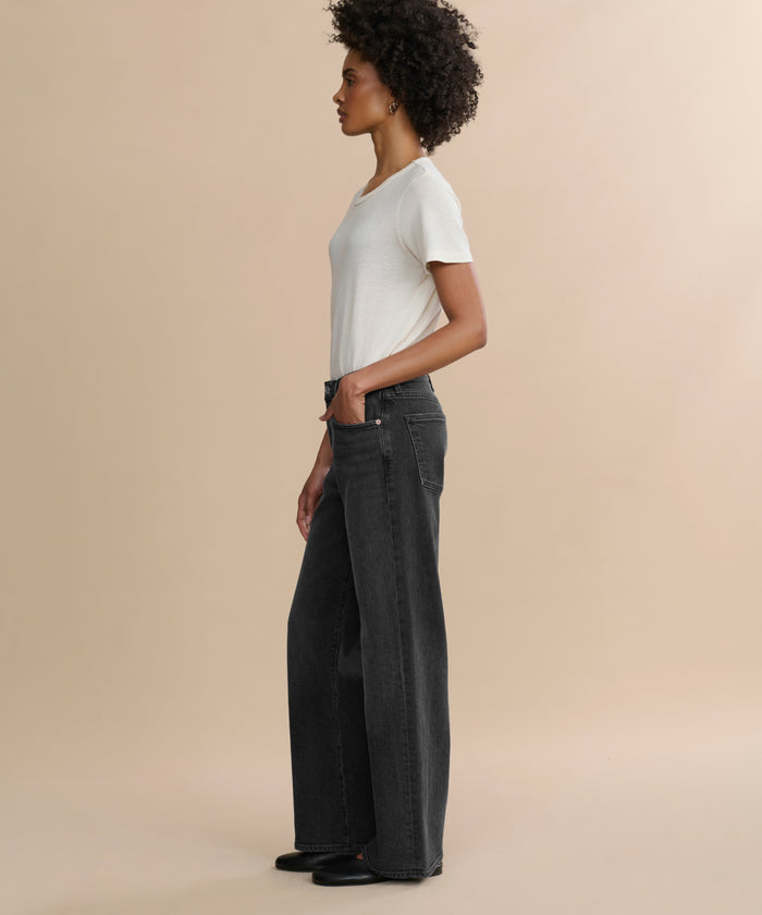 A person with curly hair stands in profile against a beige background, wearing a white T-shirt, the Harper Jean—relaxed mid-rise organic cotton jeans in a wide-leg silhouette—and black shoes, with one hand in their pocket.