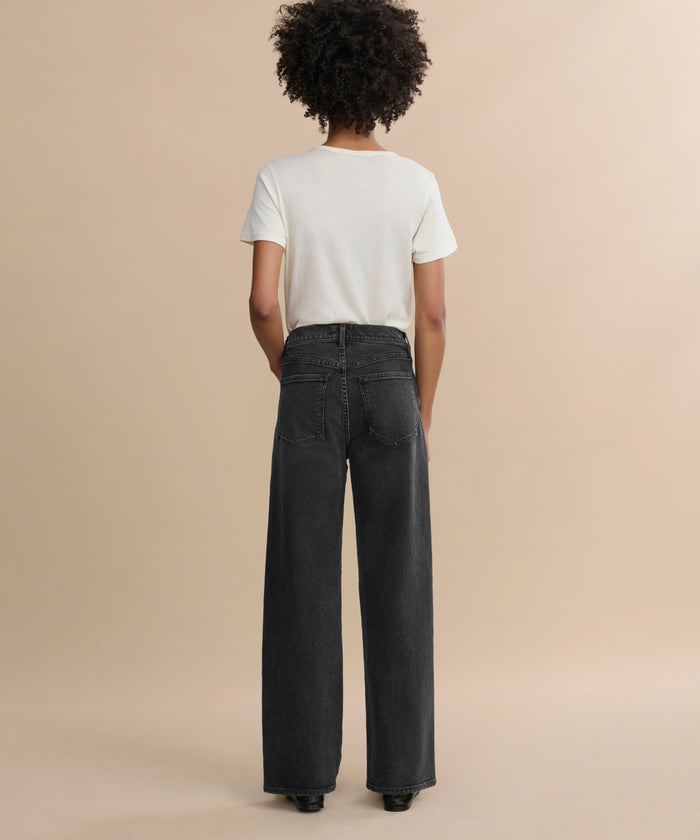 A person with curly hair faces away, wearing a white short-sleeve t-shirt and the Harper Jean—relaxed mid-rise, wide-leg jeans—against a plain beige background.
