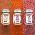 Three glass jars of Honeysuckle brand spice blends, arranged on a textured red background. The jars are labeled as 'SWEET POTATO PIE SPICE,' 'CREOLE SEASONING,' and 'HIBISCUS GINGER SUGAR.