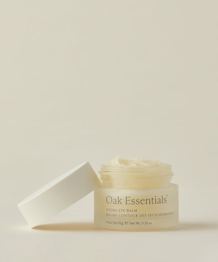 A small glass jar of Hydra Eye Balm with a white lid beside it sits open, displaying its creamy, hydrating formula that helps reduce puffiness, all set against a neutral background.