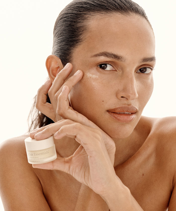 A woman with smooth skin holds a small jar of Hydra Eye Balm near her face, gently touching her cheek. With slicked-back hair and minimal makeup, she demonstrates how to reduce puffiness and dark circles against a light background.
