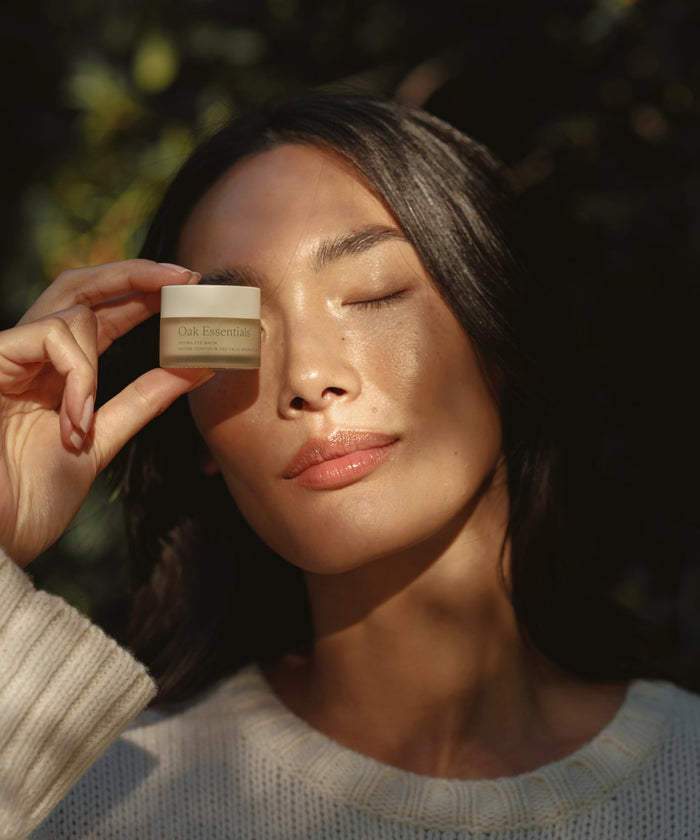 A woman with long dark hair and closed eyes, wearing a white knit sweater, gently holds a jar of Hydra Eye Balm by her face in soft sunlight, ready to reduce puffiness and dark circles.