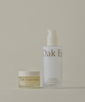 The Hydrating Double Cleanse Duo features two Oak Essentials skincare products on a beige background: a Cleansing Balm on the left and a taller Nourishing Cleansing Milk on the right, both with minimal gold-accented labels.