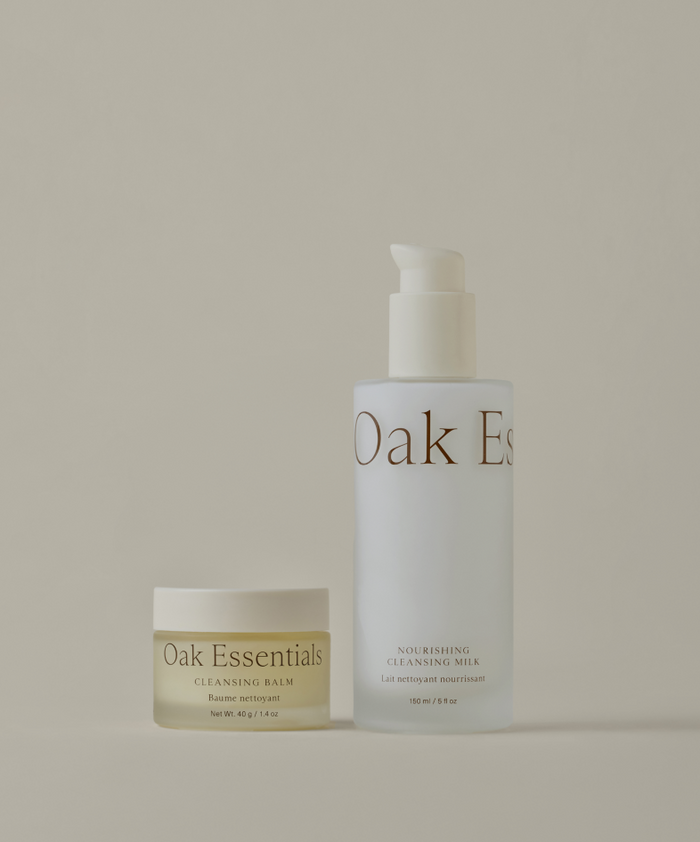 The Hydrating Double Cleanse Duo features two Oak Essentials skincare products on a beige background: a Cleansing Balm on the left and a taller Nourishing Cleansing Milk on the right, both with minimal gold-accented labels.